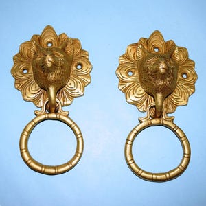 May include: A pair of antique brass-coloured door knockers. Each knocker features a fox head with a decorative floral backing and a ring-shaped handle. They are mounted on a light blue background.