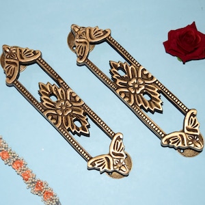 Puede incluir: Dos tiradores de puerta de latón adornados con un diseño de mariposa. Los tiradores son rectangulares con un patrón floral y una mariposa decorativa en cada extremo.