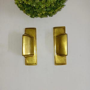 Puede incluir: Dos tiradores de armario dorados vintage. Cada tirador tiene una placa trasera rectangular con un asa cilíndrica redondeada. Los tiradores son de metal y tienen un aspecto ligeramente envejecido. El fondo es blanco.