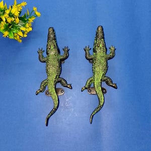 Könnte beinhalten: Zwei dekorative Türgriffe in Alligatorform, grün und schwarz. Sie sind aus Metall gefertigt und auf einer blauen Oberfläche befestigt. Die Türgriffe haben ein detailliertes Design und sind in Form von Alligatoren.