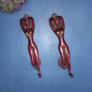 Puede incluir: Dos tiradores de puerta decorativos con forma de mujer. Los tiradores son de metal y tienen un acabado marrón rojizo.