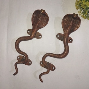 Może przedstawiać: Dwa ozdobne mosiężne klamki do drzwi w kształcie kobry z bogatymi rzeźbami. Klamki są w stylu vintage i mają ciemnobrązową patynę.