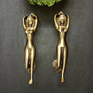May include: A pair of gold-toned metal door handles shaped like a standing female figure with arms raised above the head. The handles have a detailed design and a smooth finish.