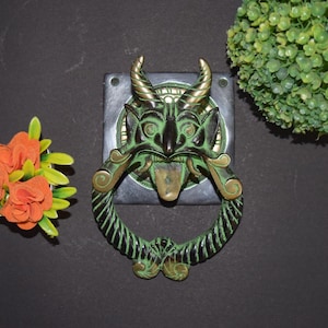 May include: A detailed, antique-style door knocker featuring a green demon face with horns and a tongue. The knocker has a ring-shaped handle and is mounted on a dark square plate. The metal has a weathered, aged appearance.