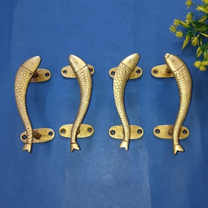 Może przedstawiać: Cztery mosiężne klamki do drzwi, każda w kształcie ryby. Klamki mają zakrzywiony kształt ze szczegółami łusek i są zamontowane na prostokątnych płytkach. Tło jest jednolicie niebieskie.
