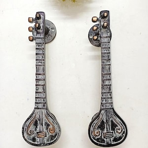 Op de afbeelding: Twee decoratieve deurklinken in de vorm van een sitar, een traditioneel Indiaas snaarinstrument. De handgrepen zijn donkergrijs met bronzen accenten en zijn op ronde platen gemonteerd.