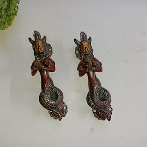 Peut inclure: Deux poignées de porte en bronze ornées en forme de femme avec une queue de serpent. La femme est en position de prière, les mains jointes. Les poignées sont de couleur brun foncé avec des accents verts et dorés.