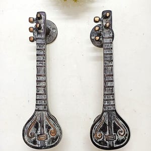 May include: Two decorative door handles shaped like sitars. The handles are made of a dark metal with bronze accents. The sitar-shaped handles have intricate detailing and are mounted on round, textured backplates.