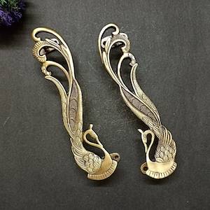 Puede incluir: Dos tiradores de puerta de metal dorado adornados con forma de pavo real con detalles intrincados. Los tiradores están sobre un fondo gris oscuro.