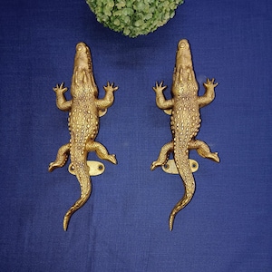 Könnte beinhalten: Zwei goldfarbene Krokodill-Türgriffe. Die Griffe sind mit strukturierten Schuppen verziert und haben ein realistisches Krokodil-Design.