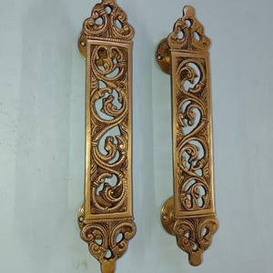 Puede incluir: Un par de tiradores de puerta de latón adornados con intrincados diseños florales. Los tiradores son de forma rectangular con una parte superior e inferior decorativas.
