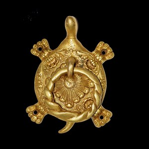 Op de afbeelding: Een gouden schildpadvormige deurklopper met ingewikkelde bloemen- en touwdetails. De klopper heeft een ringhandvat en is gemonteerd op een zwarte achtergrond. De kop en staart van de schildpad zijn zichtbaar.
