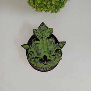 May include: A decorative, green and black metal door knocker in the shape of a stylized face. The knocker has intricate details and is mounted on a dark, circular base. The background is a plain white wall.