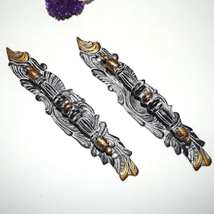 Pode incluir: Duas puxadores de armário ornamentados, prateados com detalhes dourados. Os puxadores apresentam um design detalhado de volutas e estão dispostos lado a lado. As extremidades e os detalhes centrais são dourados, adicionando contraste ao acabamento prateado. Umas flores roxas no fundo.