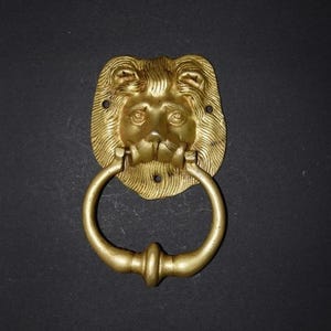 May include: A gold-colored lion head door knocker with a ring handle. The lion's face is detailed with a mane, eyes, nose, and mouth. The ring handle is attached to the lion's mouth.