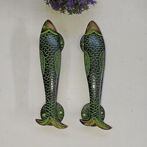 Könnte beinhalten: Zwei dekorative Türgriffe in Fischform. Die Griffe sind schwarz mit grünen und goldenen Akzenten, mit einem Schuppenmuster und Flossendetails. Die Griffe sind aus Metall gefertigt und für die Montage an einer Tür vorgesehen.