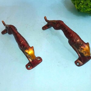 May include: Two decorative door handles in the shape of a tiger. The handles are bronze-colored with a reddish-brown body and a gold-colored head. The tiger is in a crouching position, with its tail raised.