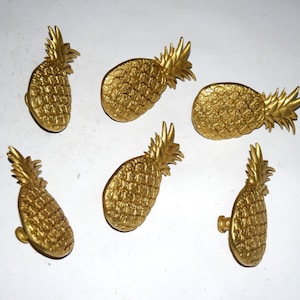 May include: Six gold-colored pineapple-shaped cabinet knobs. The knobs have a textured surface and a detailed crown. The knobs are arranged on a white surface.