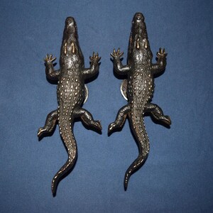 May include: Two bronze alligator-shaped door handles. The handles are detailed with textured scales and small, gold-colored accents. The alligators are positioned with their legs outstretched, giving the impression of crawling.