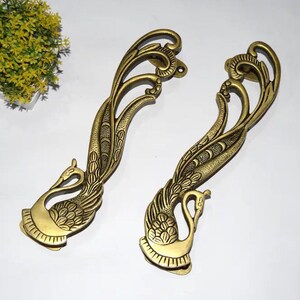 Può includere: Due maniglie antiche in ottone con un design a pavone. Le maniglie sono curve e presentano dettagli intricati, tra cui le piume e il corpo del pavone. Le maniglie sono lunghe circa 30 cm.