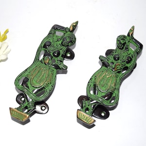 Puede incluir: Un par de tiradores de puerta de metal verde y dorado con forma de figuras danzantes. Los tiradores están detallados con intrincados tallados y tienen una base de metal negro.