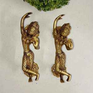 May include: Two antique gold-colored statues of dancing figures. Each statue has intricate details, including a headdress and patterned clothing. The figures are posed with one arm raised and the other bent, with a decorative base.