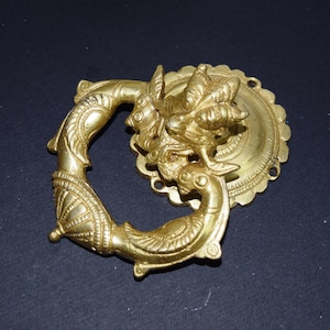 Könnte beinhalten: Ein goldfarbener, verzierter Schubladengriff mit dekorativem Design. Der Griff hat eine kreisförmige Rückplatte und einen gebogenen Griff mit detaillierten Schnitzereien von Fabelwesen und Mustern. Das Gesamtdesign ist symmetrisch und antik inspiriert.
