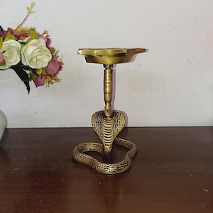 以下が含まれることがあります： 丸い天板が付いたコブラの形をした金色真鍮製のオイルランプスタンド。スタンドは木製の表面の上に置かれています。