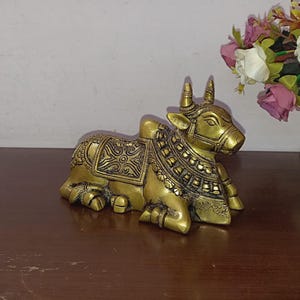 May include: A gold-colored brass sculpture of a reclining cow with intricate details. The cow has a decorative collar and a patterned design on its back.