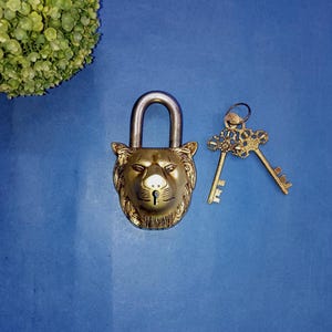 May include: A brass padlock with a lion's head design, featuring a silver shackle and two decorative keys. The lock is gold-colored, with intricate details on the lion's face. The keys have ornate floral patterns. The background is a solid blue.