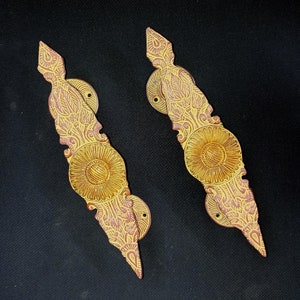 Könnte beinhalten: Zwei verzierte Türklinken mit goldener und rosafarbener Oberfläche. Die Griffe sind in Form eines langen, spitzen Blattes mit einem dekorativen Blumendesign geformt. Die Mitte jedes Griffs weist ein rundes, erhabenes Design mit goldener Oberfläche auf.
