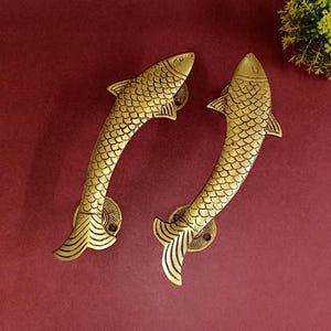 Op de afbeelding: Een paar goudkleurige messing deurgrepen in de vorm van vissen. De grepen hebben een gedetailleerd, schubbig ontwerp en zijn gebogen om comfortabel in de hand te passen.