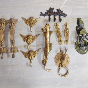 May include: A collection of brass hardware including two long vertical handles with figure motifs, three angel-shaped wall hooks, a dark metal frog hook, a leopard-shaped handle, two small rectangular pulls, a deer head door knocker, and a bird knocker.