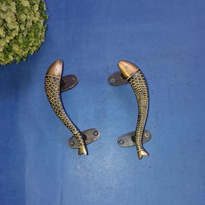 May include: Two brass door handles shaped like fish with a black finish and gold accents. The handles are mounted on a black metal plate.