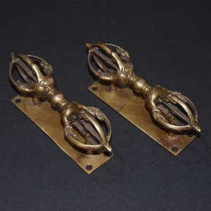 Peut inclure: Deux sceptres vajra en laiton antique, chacun avec une poignée détaillée et une tête stylisée à plusieurs pointes. Les sceptres sont montés sur des plaques rectangulaires en laiton avec trois trous, sur fond sombre.