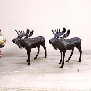 May include: Two bronze-colored moose figurines with detailed antlers stand side-by-side. The moose are posed in a standing position, with their legs slightly bent. The figurines are placed on a marble-like surface, with a bouquet of flowers in the background.