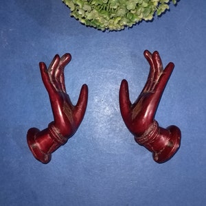Könnte beinhalten: Zwei rote Holz Hände mit detaillierten Schnitzereien, jede mit einer nach oben gerichteten, erhobenen Handfläche. Die Hände sind an einer Basis befestigt und scheinen dekorativ zu sein.