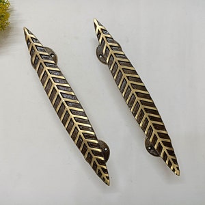 Peut inclure: Deux poignées de porte en laiton avec un motif de feuille. Les poignées sont longues et étroites, avec une extrémité pointue. Le motif de la feuille est complexe et détaillé, avec un motif en chevron.