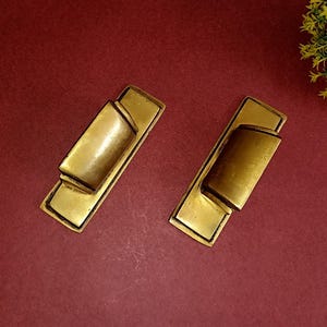 Puede incluir: Dos tiradores de cajón de metal dorado con forma rectangular y un centro hundido. Los tiradores tienen un acabado ligeramente envejecido.