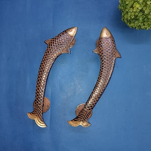 May include: Two gold-coloured fish-shaped door handles with brown scales. The handles are designed to be mounted on a door.