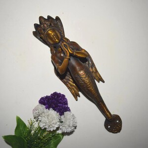 Può includere: Una maniglia per porta a forma di sirena color bronzo con corona e ali dettagliate. La sirena è in posizione di preghiera. La maniglia è montata su una superficie bianca, con un piccolo bouquet di fiori viola e bianchi.