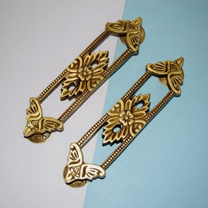 May include: Two ornate, gold-colored drawer pulls with floral and leaf designs. Each pull has a rectangular frame with decorative corners and a central floral motif. The pulls are set against a light blue and white background.