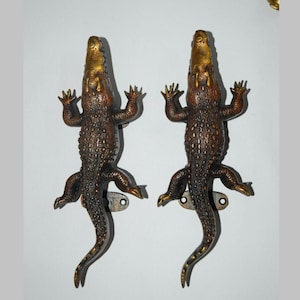 May include: Two bronze-colored alligator-shaped door handles. The handles are detailed with textured scales and have gold-colored accents on the head and feet. Each handle has a mounting plate with a single screw hole.