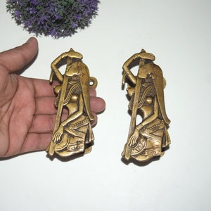 Puede incluir: Dos tiradores de puerta de metal dorado con forma de mujeres con ropa tradicional india. Los tiradores están detallados con intrincados tallados y tienen un acabado ligeramente envejecido.