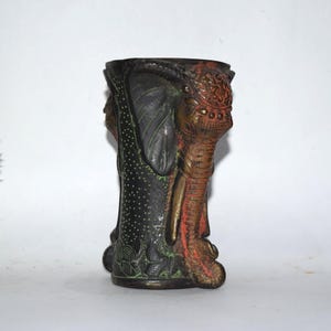 Puede incluir: Un jarrón de bronce con diseño de elefante. El jarrón presenta cabezas de elefante en cada lado, con detalles intrincados y acentos rojos, verdes y dorados. El cuerpo del jarrón tiene un patrón de puntos.