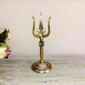 May include: A brass trident sculpture with a three-pronged head and a long, slender handle. The sculpture has a detailed design and a polished finish. The base is round with concentric circles. The trident is a symbol of power and authority.