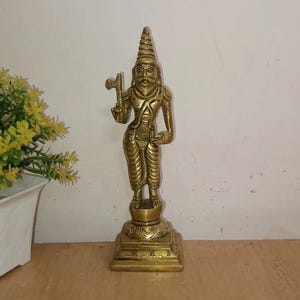May include: A brass statue of a male figure, standing on a square base. The figure is adorned with detailed clothing and a tall, conical headdress. He holds a small axe in one hand. The statue is a warm, golden color.