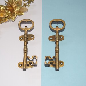 Może przedstawiać: Dwa mosiężne uchwyty do drzwi w kształcie klucza z ozdobnym wzorem. Uchwyty są zamontowane na białym i niebieskim tle.