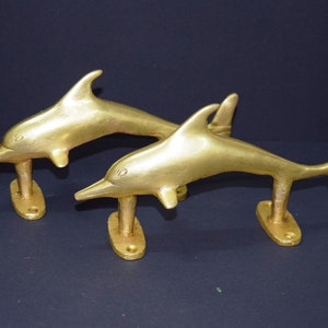 Könnte beinhalten: Zwei goldfarbene Delfin-Türgriffe. Die Delfine sind nach vorne gerichtet, mit nach oben gekrümmten Schwänzen. Die Griffe sind auf einem rechteckigen Sockel montiert.