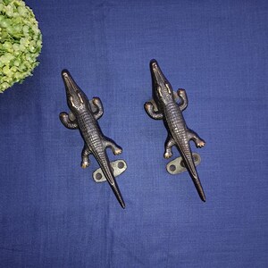 May include: Two black metal alligator door handles with a textured finish. The handles are mounted on a blue surface.